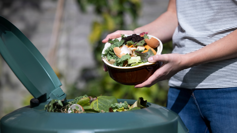 Person holding bowl filled with discarded food scraps over compost bin outdoors.