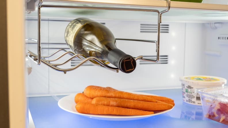 Metallic wire wine rack hangs from the middle shelf of the fridge, suspending a bottle of white wine over a bunch of carrots.