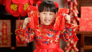 Little girl joyfully holding two red envelopes with festive Lunar New Year decorations behind her.