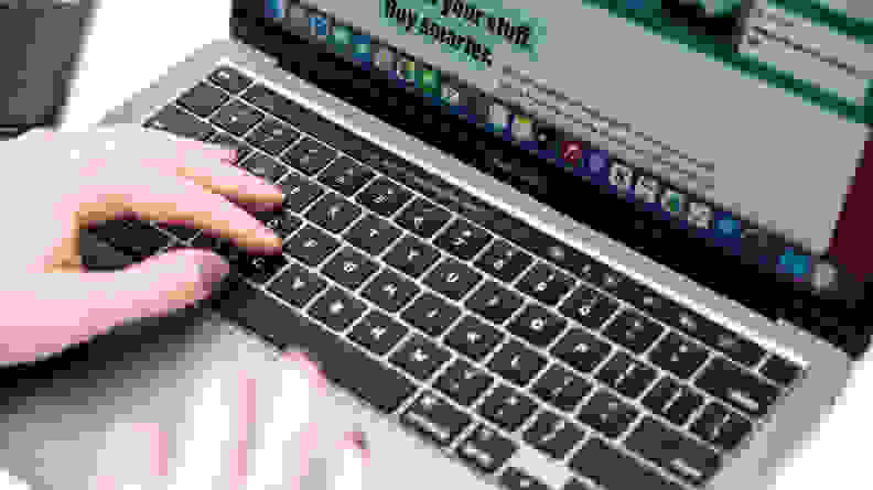 An over-the-shoulder photograph of someone using a MacBook Pro. A small touchscreen toolbar above the keyboard serves as a search bar, volume controls, and so forth.