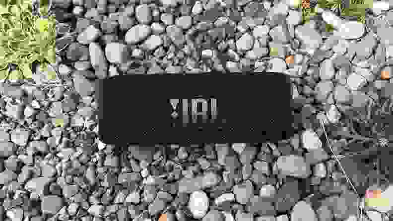 A black, cylindrical Bluetooth speaker sits in the gravel with a JBL logo showing.