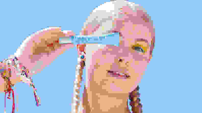 A person with long blond hair in braids on either side of their head holds up a blue squeeze tube in front of their eye while having yellow dots all around their face.