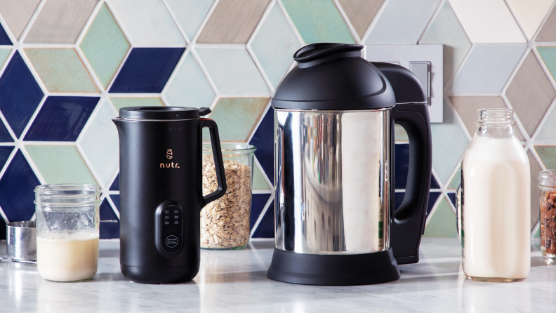 The Nutr plant-based milk maker next to the Almond Cow plant-based milk maker on granite countertop in front of tiled wall and 3 clear jars with snacks inside.