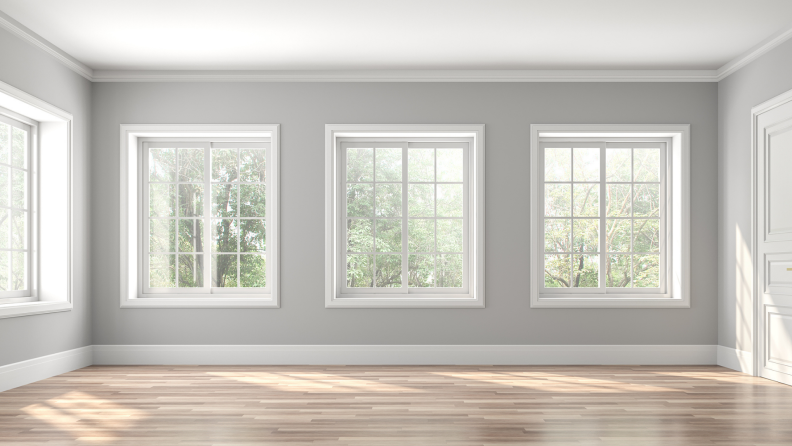 An empty but beautiful room features light-gray walls, several large windows with white-painted trim, and natural hardwood.