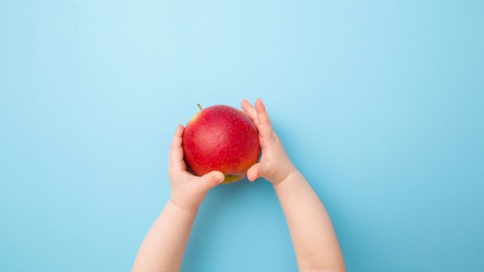 A toddlers hands extended holding a red apple against a light blue background.