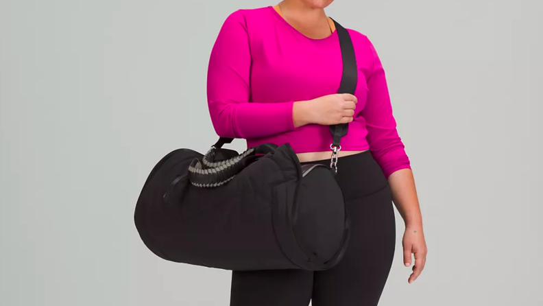 An image of the lululemon Barrel Duffel Bag slung over the shoulder of a model wearing a bright pink cropped shirt and black leggings.