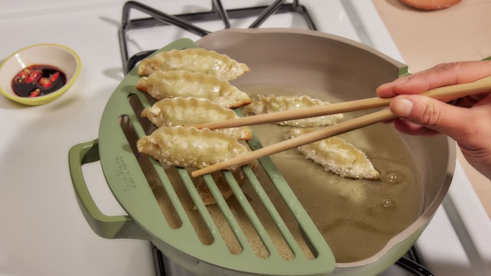 An Always Pan filled with pan-frying dumplings, Fry Deck attached on the left side of the pan with some dumplings placed there. Chopsticks holding one of the dumplings.