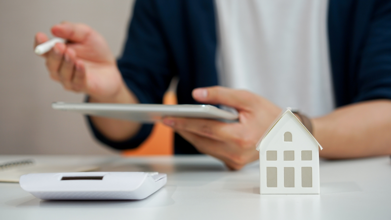 A person working on a calculator with a tiny model house on the table in front of them.