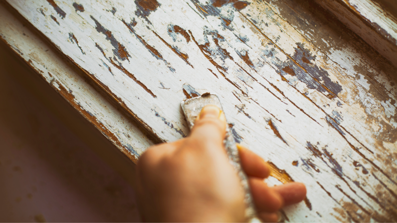 A person strips a layer of old paint from a wooden window frame.