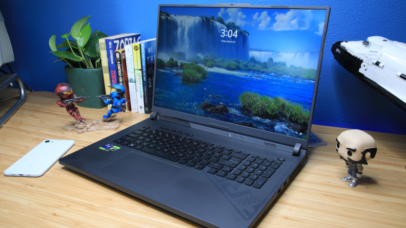 The black Asus ROG Strix G18 gaming laptop sitting on top of wooden desk next to toy trinkets, books and a potted plant.