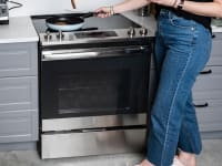A person wearing jeans stands in front of an electric range cooking food in a pan on the stovetop.