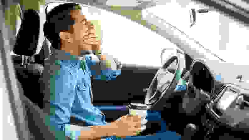 Person in car yawning while holding coffee cup.