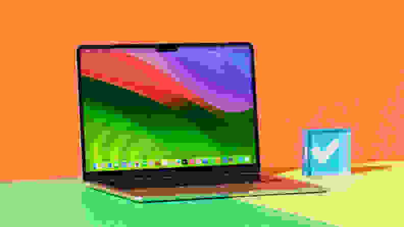 The black laptop sits on a green square on a desk, in front of an orange background