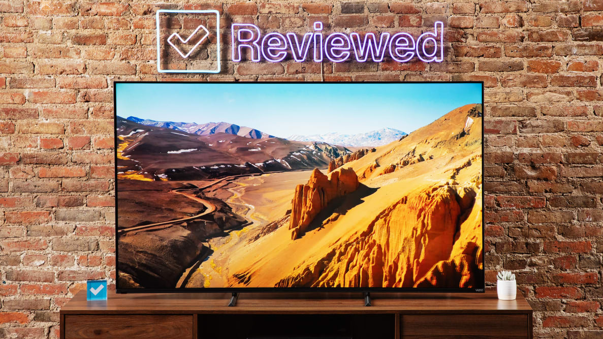 The Vizio M-Series Quantum X TV sitting on a wooden credenza in front of a brick background, displaying an outdoor scene.