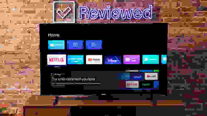 The Hisense U8H on a wooden credenza displaying Google OS with a brick wall in the background and a Reviewed neon sign.
