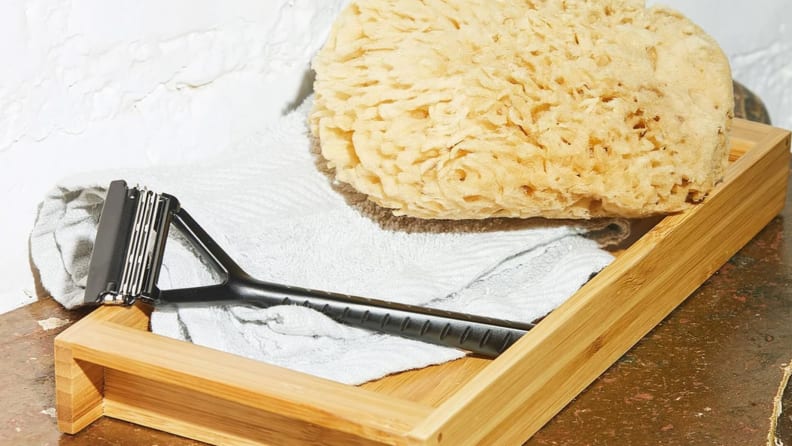A dark gray stainless steel razor from Leave Shave sits on top of a white towel with a loofa next to it.