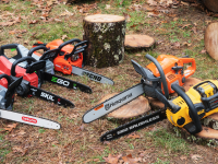 Six chainsaws sitting on the ground outside near a tree stump