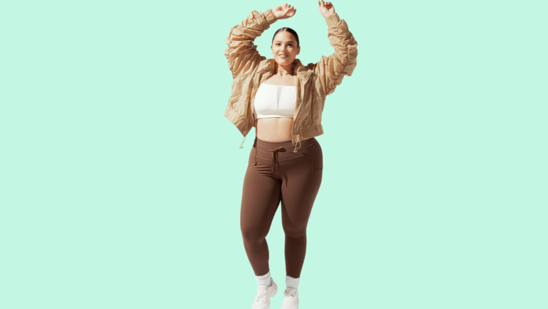 A model wearing brown tights, a white sports bra, and a tan jacket.