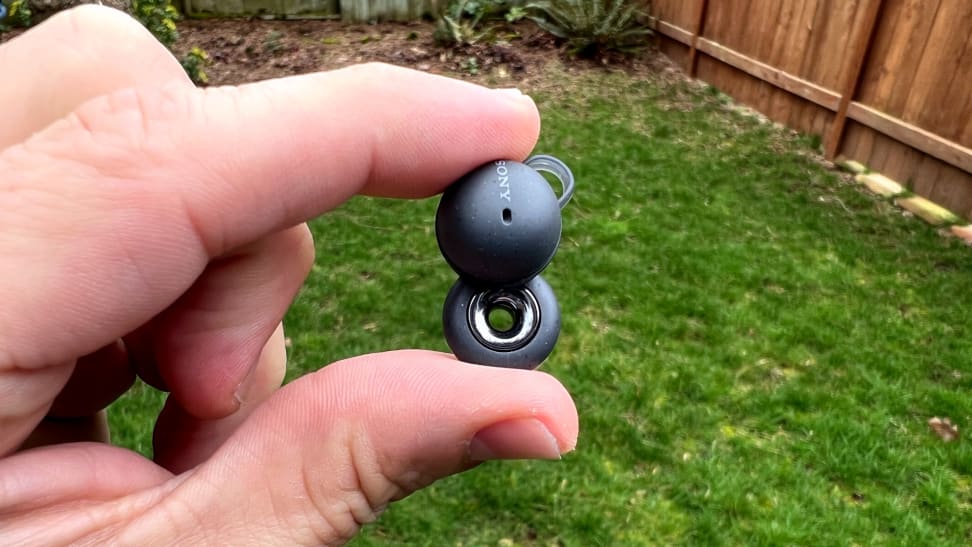 A hand holds a double-looped gray earbud in front of a grass background with a cedar fence in view.