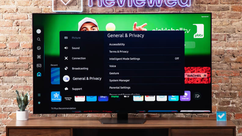 Samsung QN90D Mini-LED TV with menu bar on screen atop a wooden TV stand below Reviewed neon sign in front of brick wall indoors.
