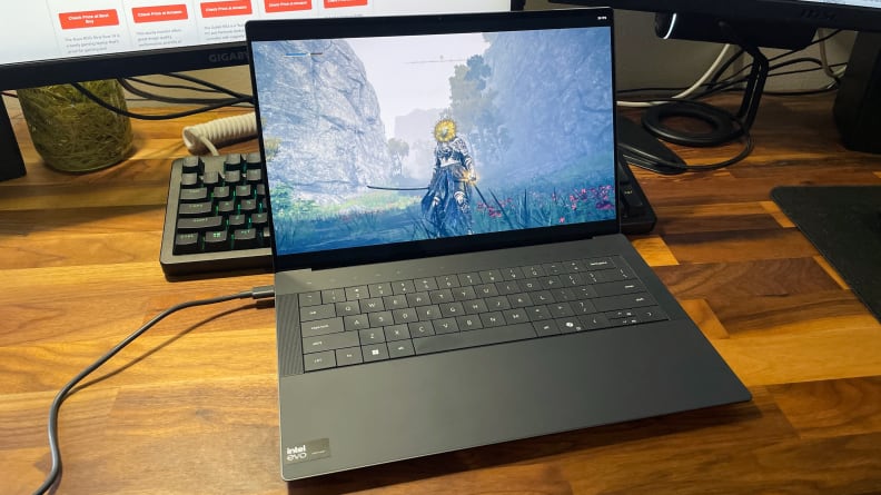The Dell XPS 14 laptop opened to display a game on screen on a wooden desk in front of a desktop computer monitor.