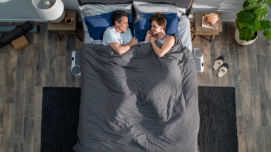A couple lies in bed smiling at each other under a shared blanket, with Chilipad Dock Pro units visible on both sides of the bed.