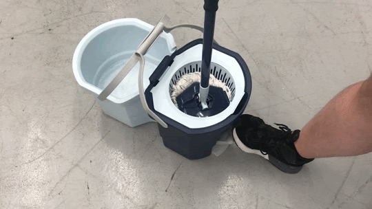 A foot in sneakers presses the mop bucket spin pedal, causing it and the adjoining bucket to wobble.