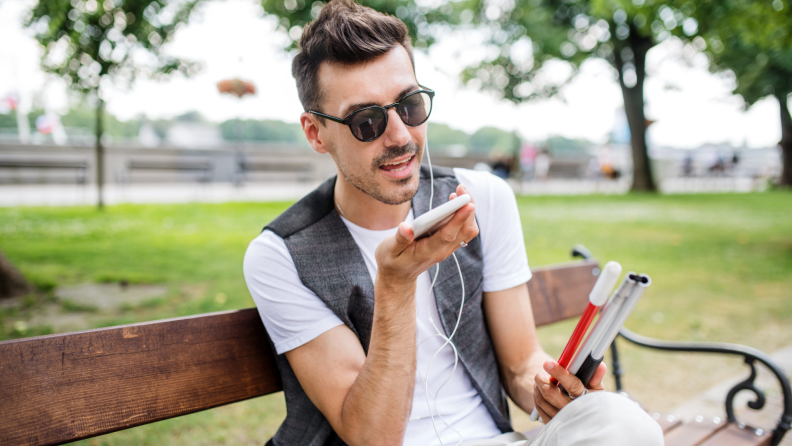 Blind person sitting on park bench outdoors using smartphone.