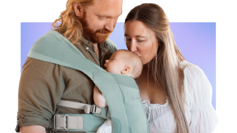 parents hold their baby in an Ergo carrier. The dad wears the baby while the mom kissed them.