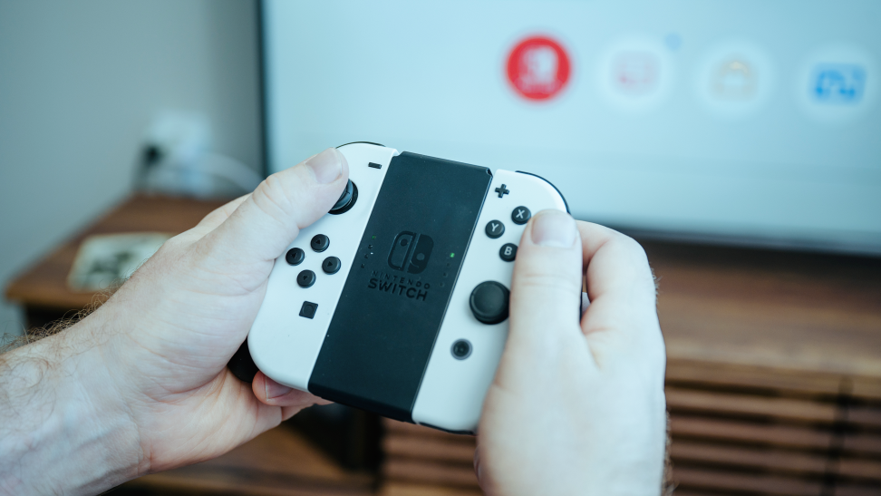 Close-up of the Nintendo Switch OLED controller with white Joy-Con attached on either side. The Switch menu UI is displayed on the TV in the background.