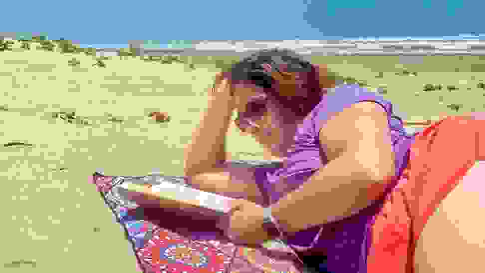 A woman reads a book while laying on a beach towel on the sand.
