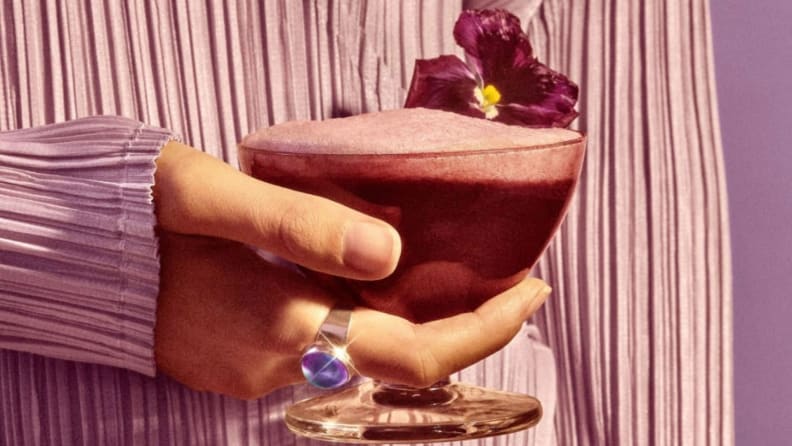 A hand holds a violet-hued cocktail garnished with a tropical flower.