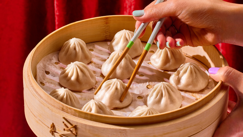 Person using chopsticks to pick up a dumpling from a bamboo dumpling steaming basket