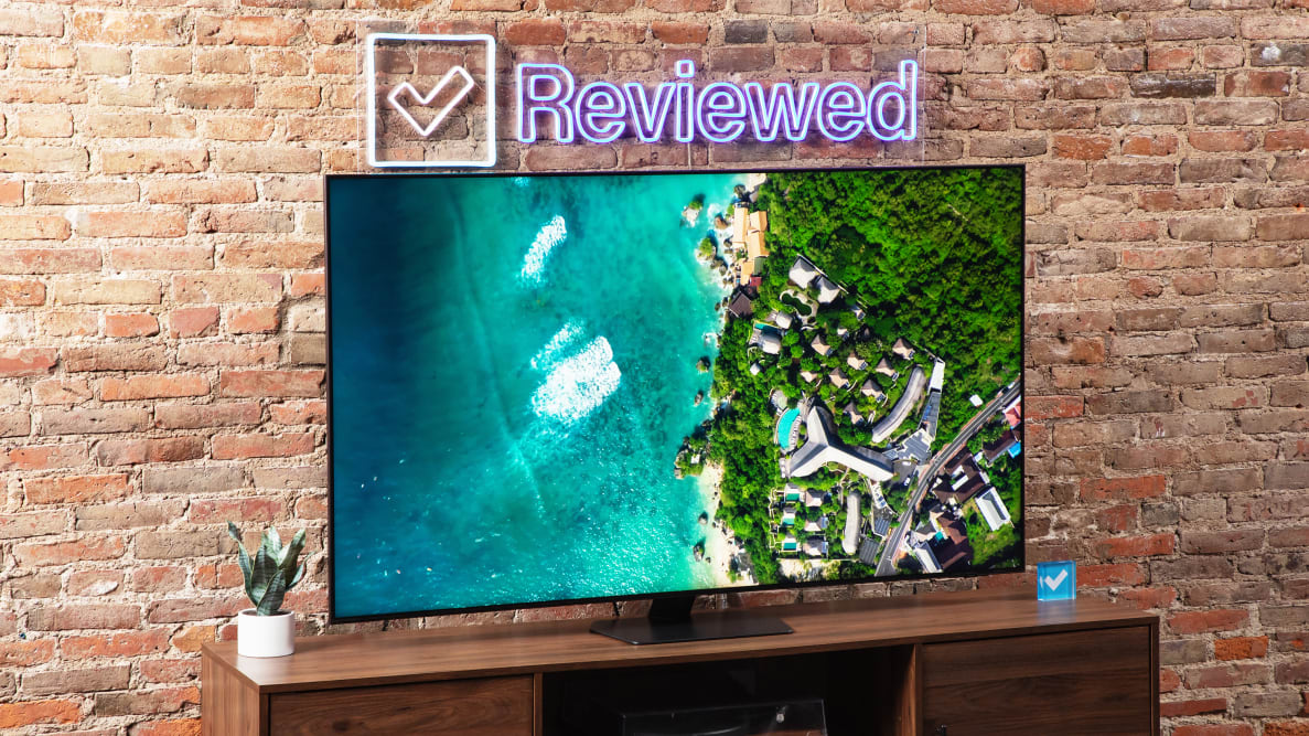 Samsung QN90D Mini-LED TV on top of wooden TV stand below Reviewed neon sign in front of brick wall indoors.