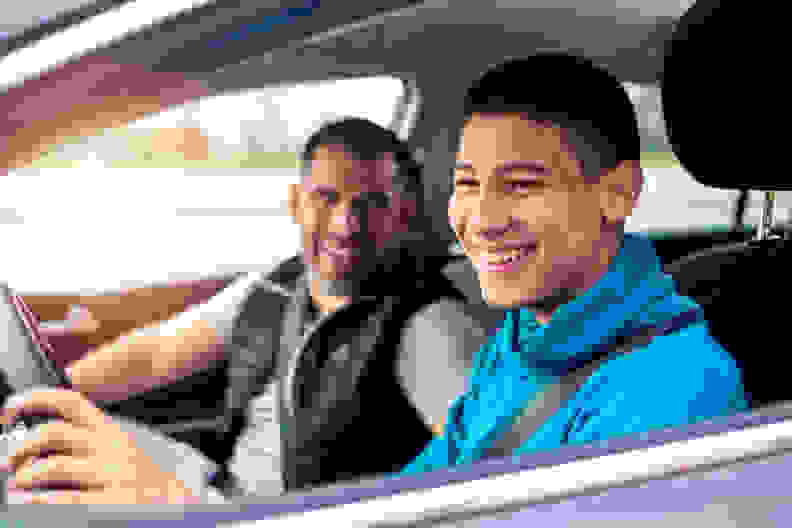 A teenage boy sitting behind the steering wheel of a car and listening to his father's instructions as he drives.