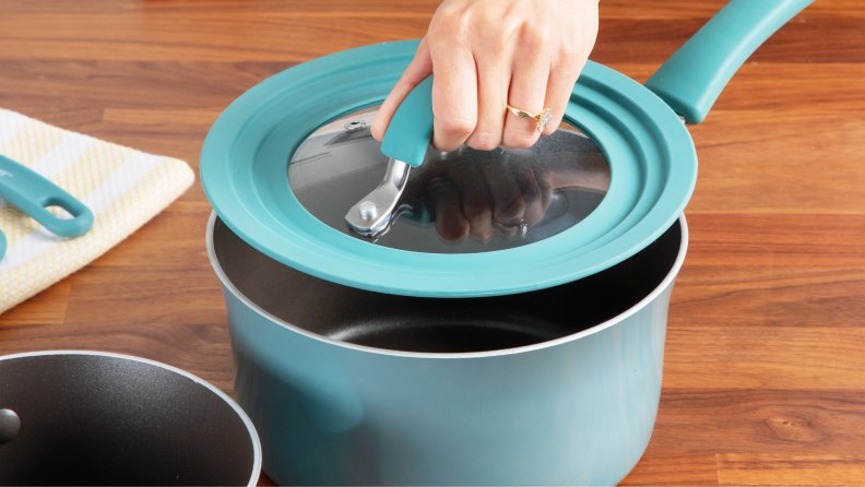 Person placing matching lid on top of small pot which sits on wooden countertop surface.
