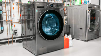 The LG WM6998HBA, a silver front load washing machine and dryer combination sits against a wall covered with pipes.