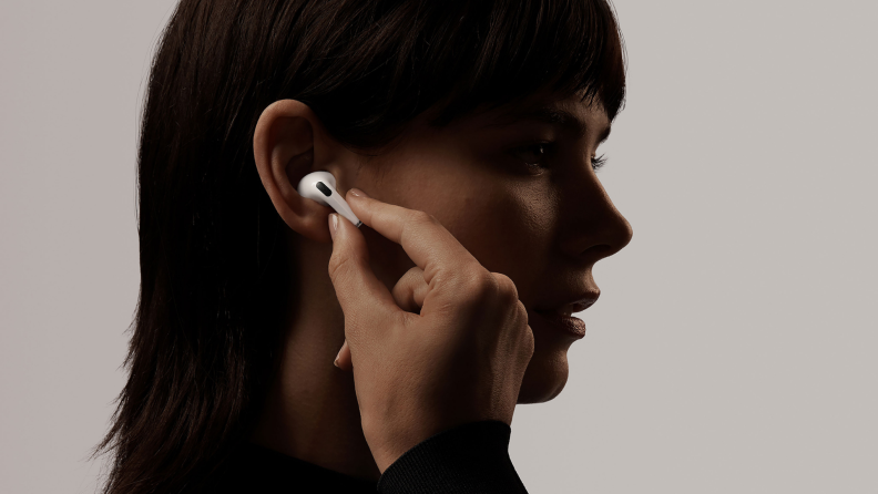 A woman with shadow on her face putting an AirPod into her ear against a grey background.