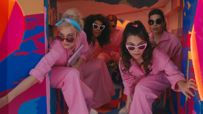 A film still from the Barbie movie in which several individuals are coming out of the back of a van, each wearing a pink jumpsuit and sunglasses.