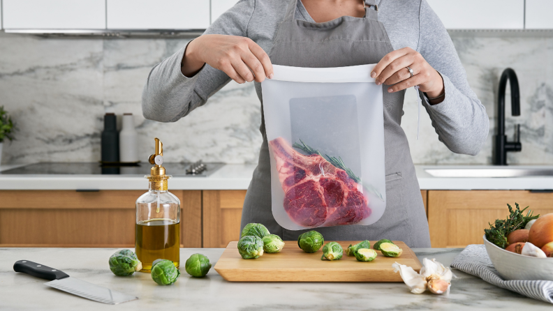 In a spacious kitchen, a person is lifting a silicone bag with a large piece of steak in it. There's a bottle of olive oil, some Brussels sprouts, a cutting board, and some onions in front of the person.