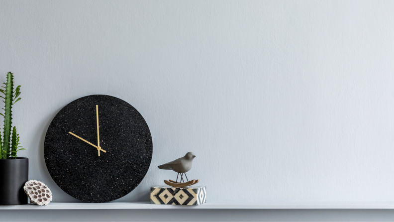 A cactus, black clock, and bird figurine sit on a shelf against a grey wall.