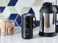 The Nutr plant-based milk maker next to the Almond Cow plant-based milk maker on granite countertop in front of tiled wall and 3 clear jars with snacks inside.
