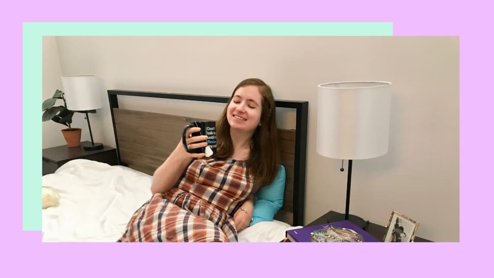 A woman sitting on a bed holding a mug with a teabag string hanging out of it.