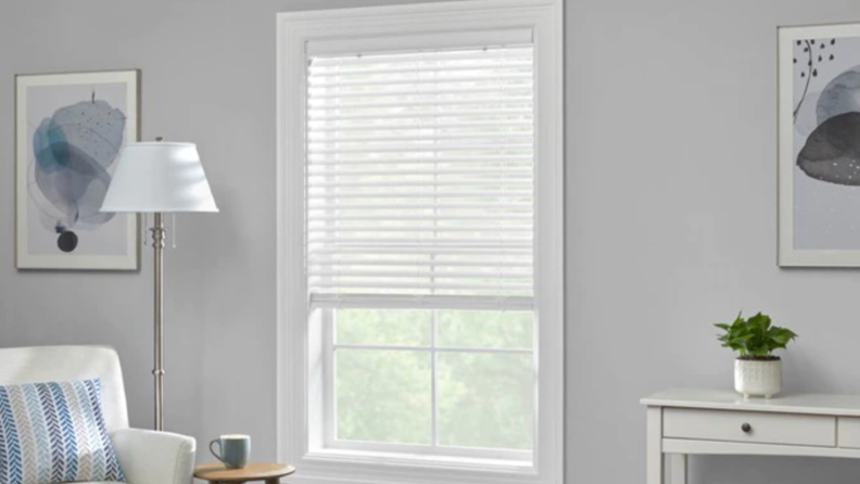 An image of a set of inside, slatted blinds in white set into a white window frame in a gray living room space.