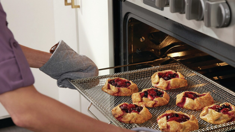 A mitten-wearing person is taking a tray of galettes with berry jam filling out of the oven.