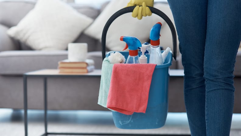 Person holding bucket handle with cleaning supplies inside in living room.