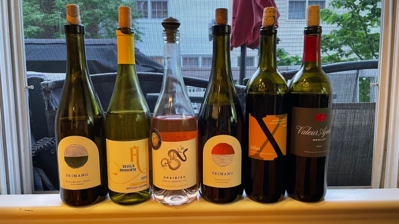 Six bottles of corked wine (a variety of whites, reds, and rosé) on a window sill in front of an open window.