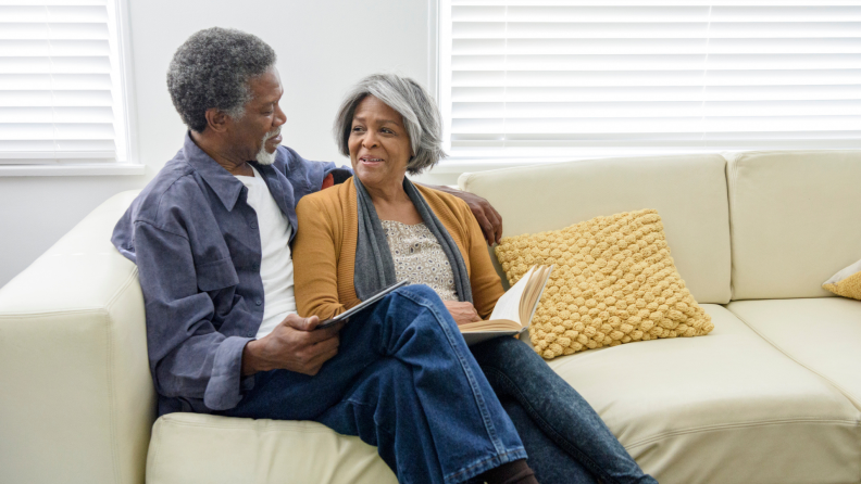 Older couple sitting on couch together, one holding a tablet and the other reading a book, facing each other smiling