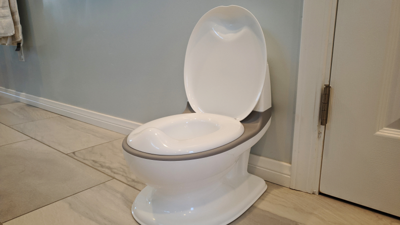 A child's potty seat with the lid open sitting on a bathroom tile floor.