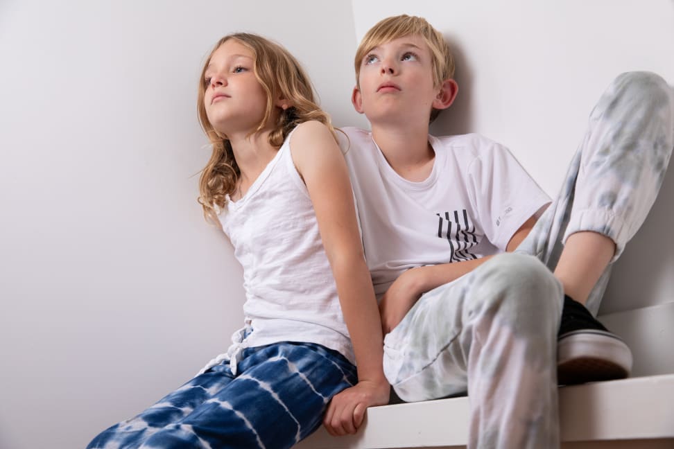 A boy and girl, both wearing white shirts and tie dye sweatpants, sit against a wall